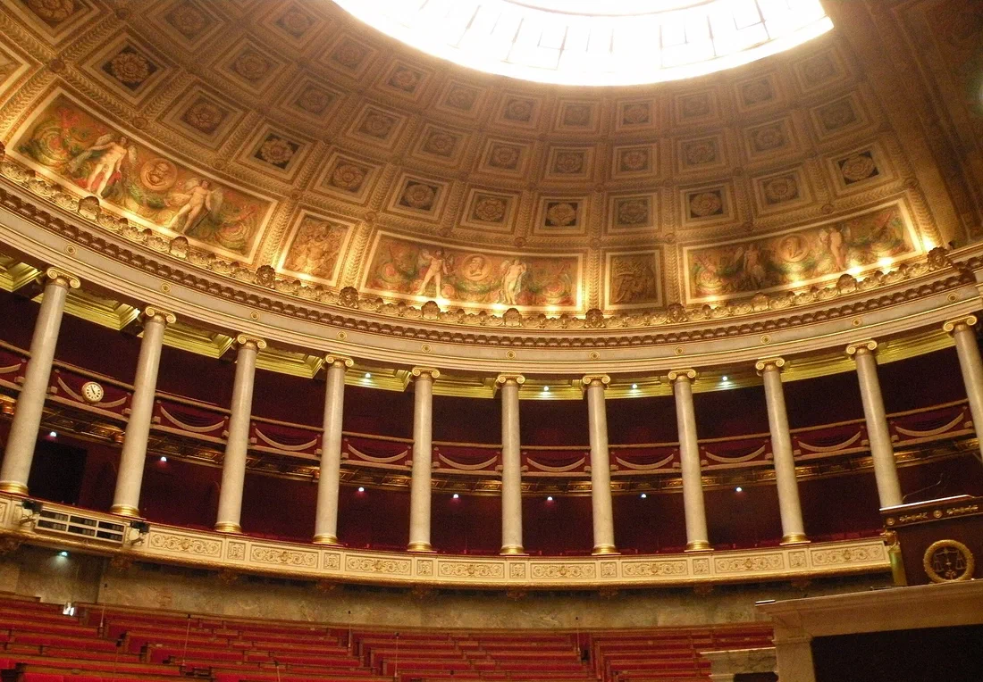 L'hémicycle de l'Assemblée nationale, vide : colonnes, tribunes drapées de rouge, coupole à caissons et rangées de sièges en bas de l'image