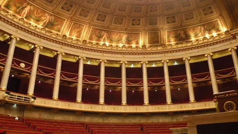 L'hémicycle de l'Assemblée nationale, vide : colonnes, tribunes drapées de rouge, coupole à caissons et rangées de sièges en bas de l'image