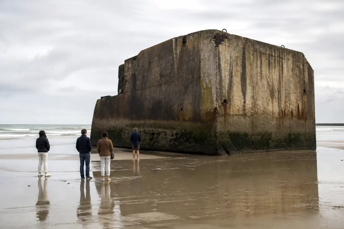 Illustration générée par IA de visiteurs anonymes contemplant un caisson de béton rouillé d&rsquo;un port artificiel, échoué sur une plage normande à marée basse